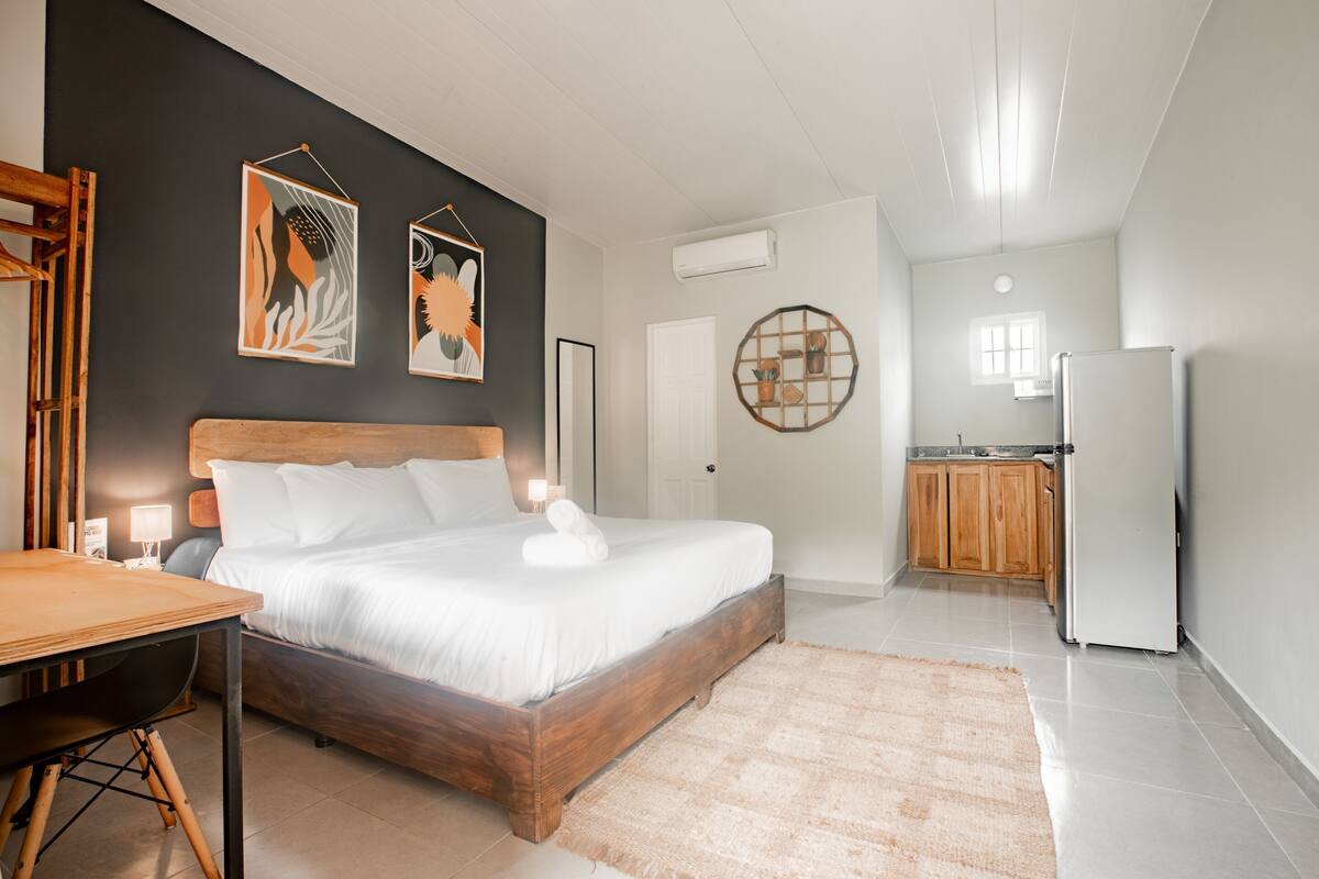 A bedroom with a wooden bed, a small table, and a kitchen area in the background.