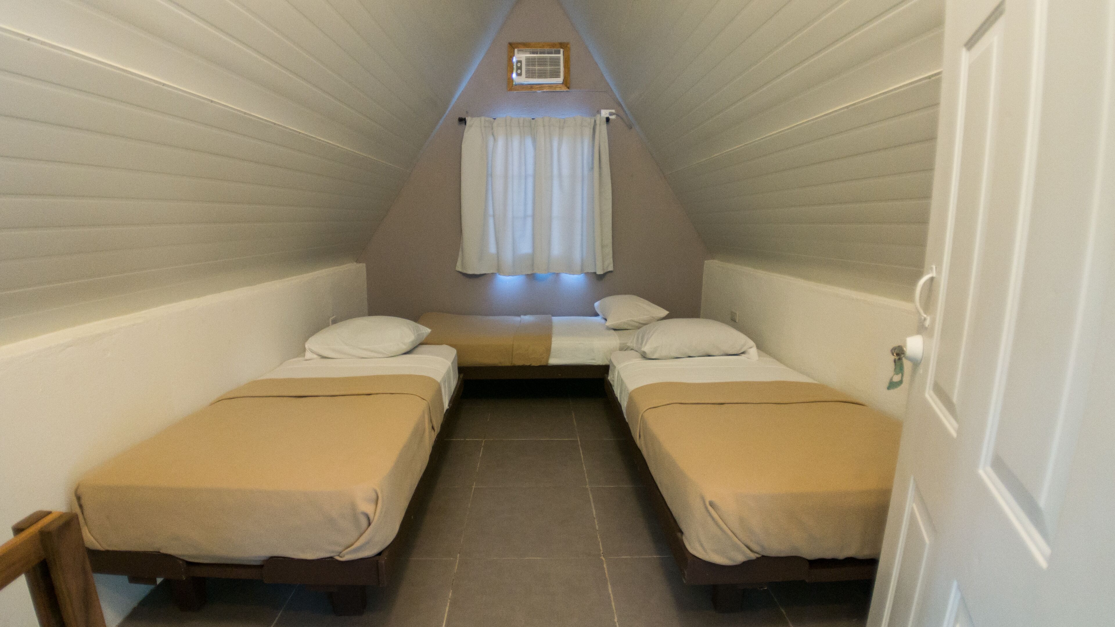 Una habitación pequeña y triangular con dos camas individuales, una ventana con cortinas y una rejilla en el techo.