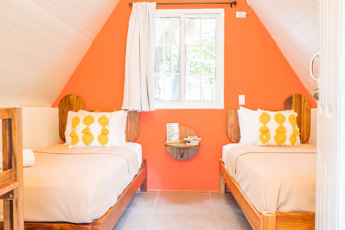 A room with two beds, a window with curtains, a small table, and a wall-mounted shelf.