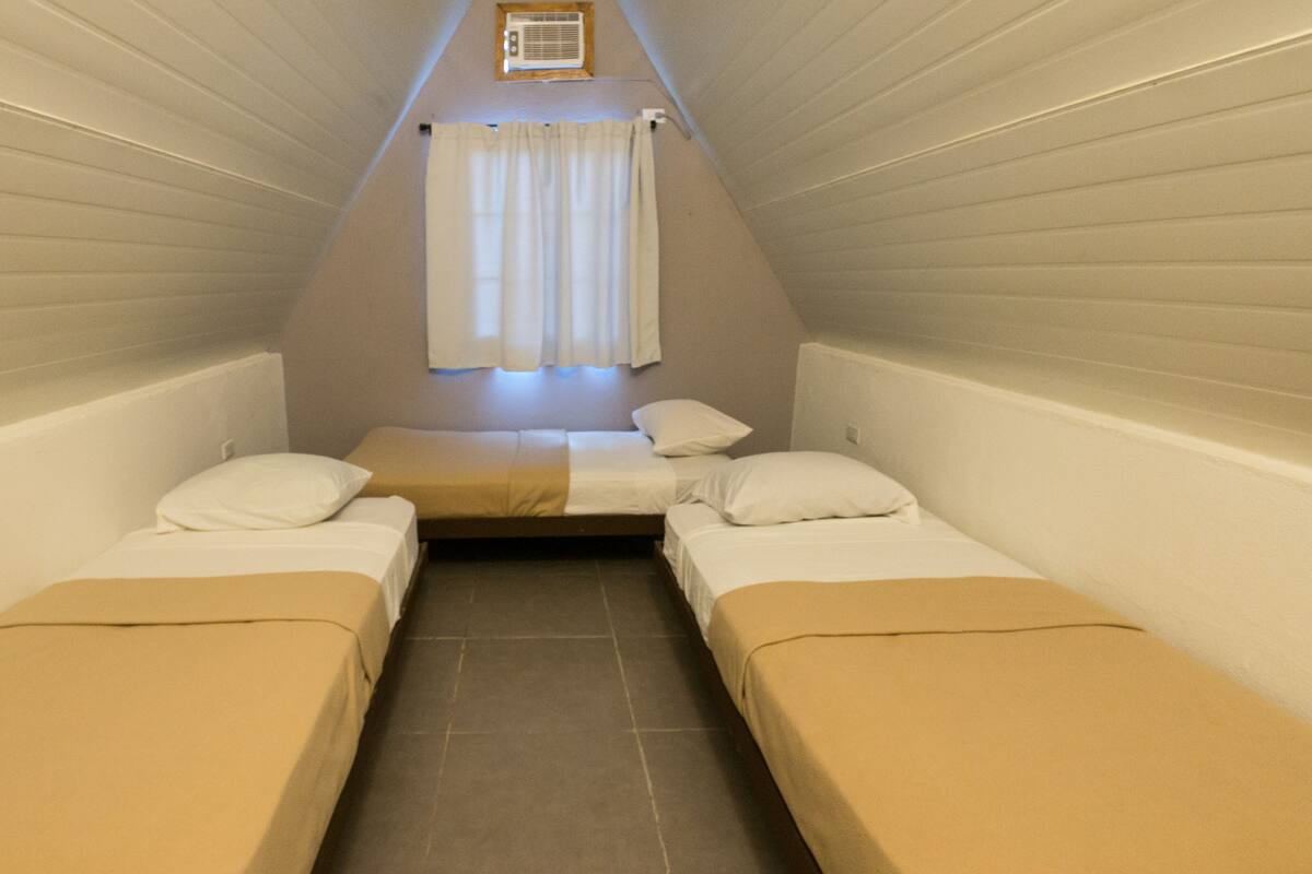 A small, triangular room with two single beds, a window with curtains, and an air conditioning unit on the wall.
