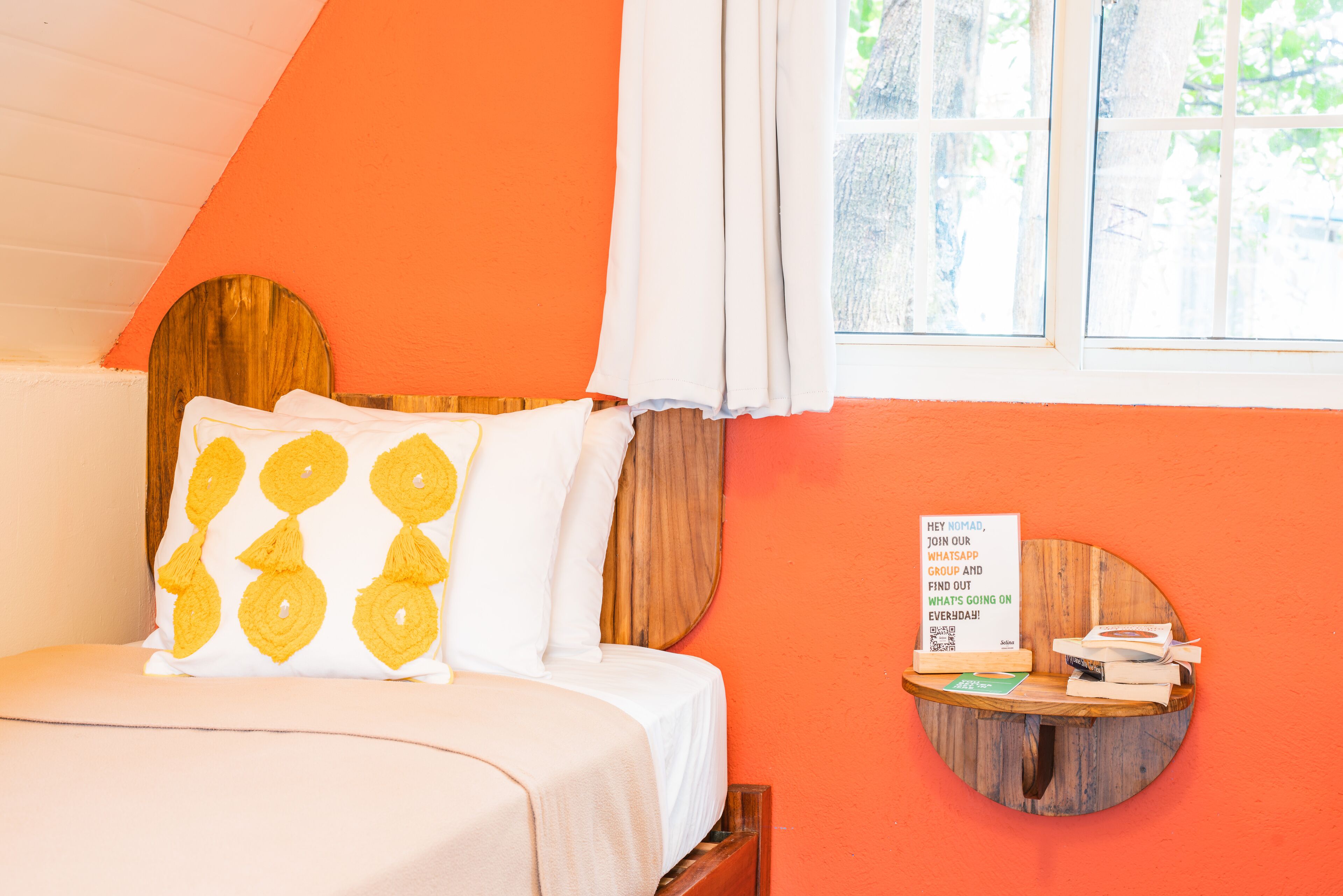 Una cama con cabecera de madera, almohada blanca y amarilla, y un pequeño estante de madera con libros y un letrero.