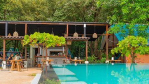 An outdoor pool area with wooden tables and chairs, surrounded by lush greenery and a covered seating area with hanging lanterns. - Selina River Venao (Las Escobas del Venado)