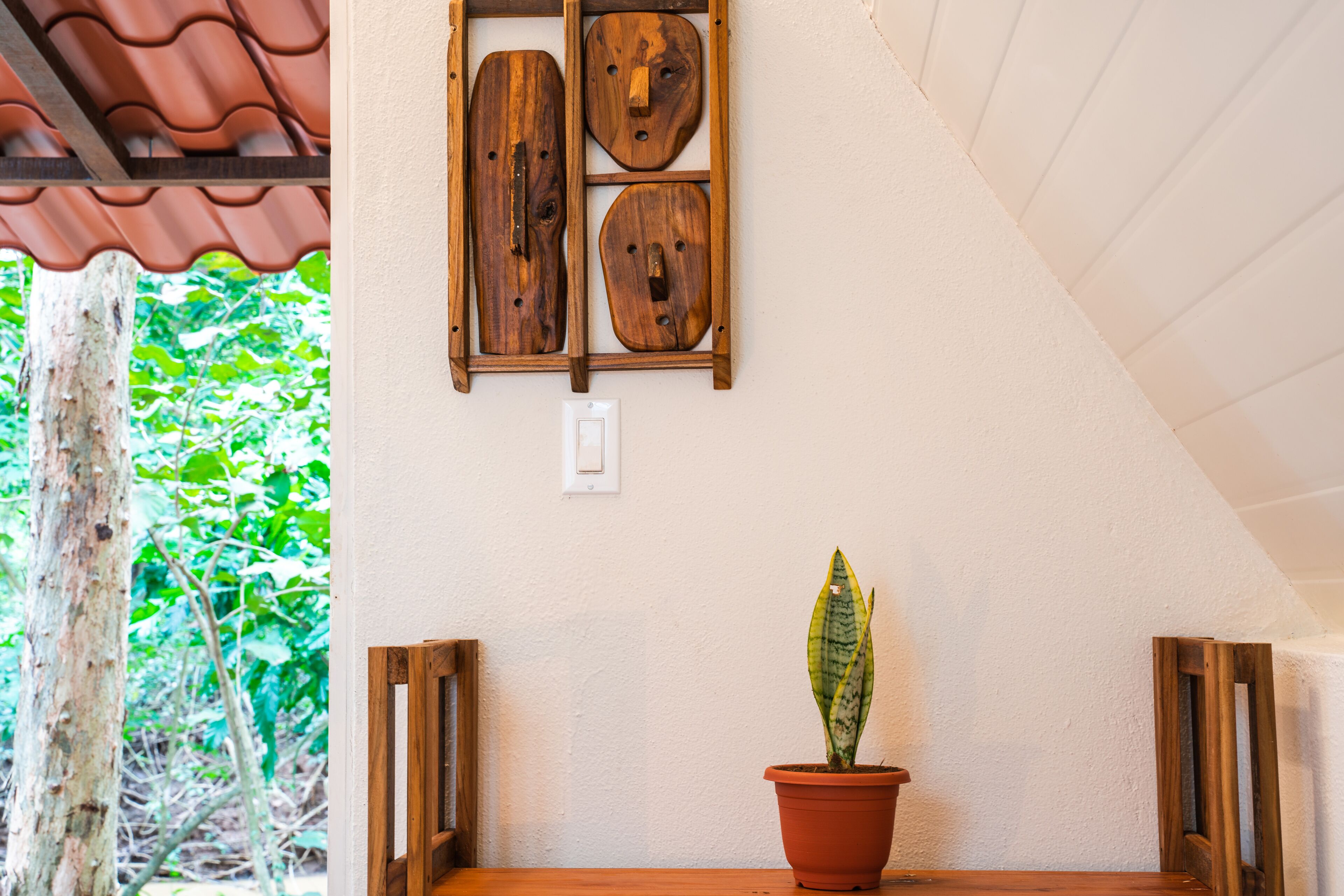 Una planta en maceta sobre una mesa de madera, con una pared blanca y un estante de madera con cuatro objetos de madera.