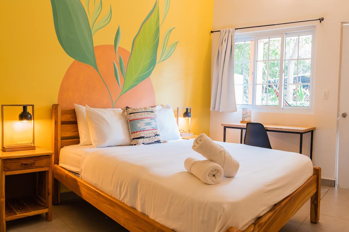 A neatly made bed with white linens and a rolled towel, a wooden nightstand with a lamp, a desk with a chair, and a window with curtains.
