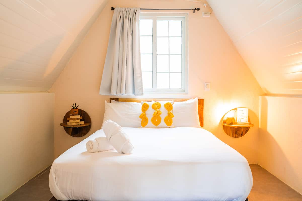 A bedroom with a large bed, a window with curtains, a bedside table with a lamp, and a wall-mounted shelf with a plant.