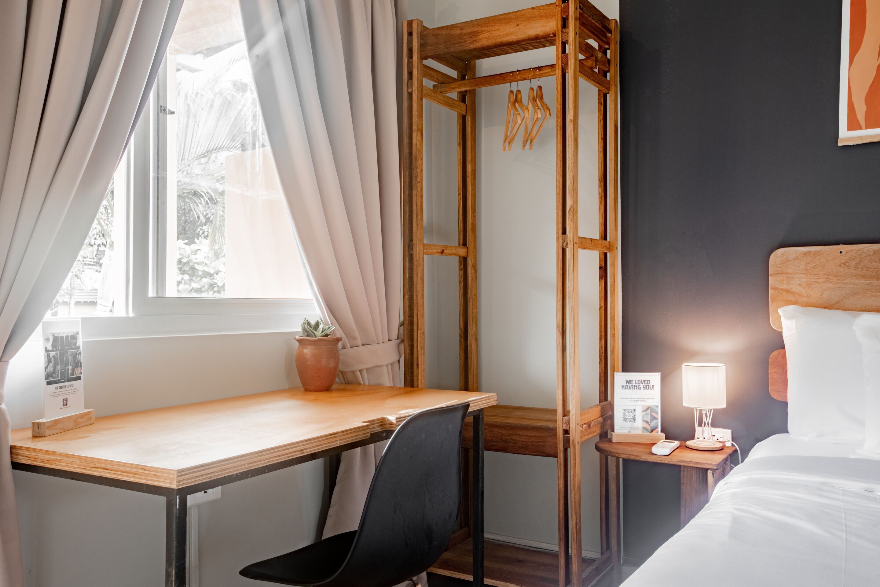 Une chambre d’hôtel avec un bureau en bois, une chaise noire, un lit avec une literie blanche, une table de chevet avec une lampe et une étagère murale agrémentée d’une plante en pot.