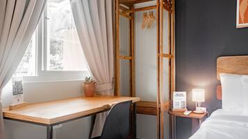 A hotel room with a wooden desk, a black chair, a bed with white bedding, a bedside table with a lamp, and a wall-mounted shelf with a potted plant.