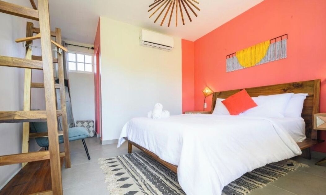 A bedroom with a wooden ladder, a bed with white bedding and a red pillow, a wall-mounted air conditioner, and a patterned rug.