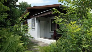A white house with a brown roof, surrounded by lush greenery and a stone pathway leading to the entrance.
