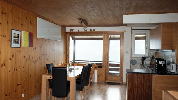 A wooden-paneled room with a dining table and chairs, a kitchen area with a coffee machine, and a large window.