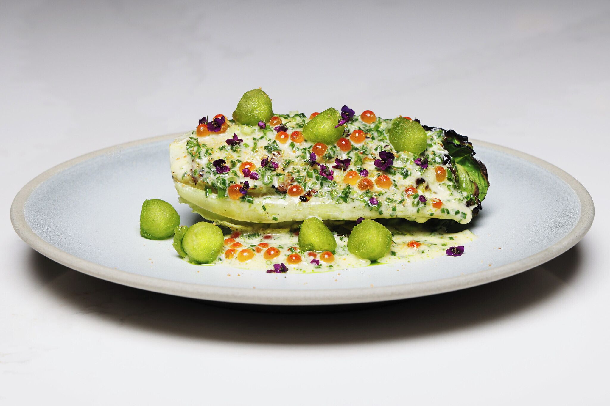 Une assiette contenant un plat, peut-être un filet de poisson, agrémenté de sphères vertes et de fleurs violettes.