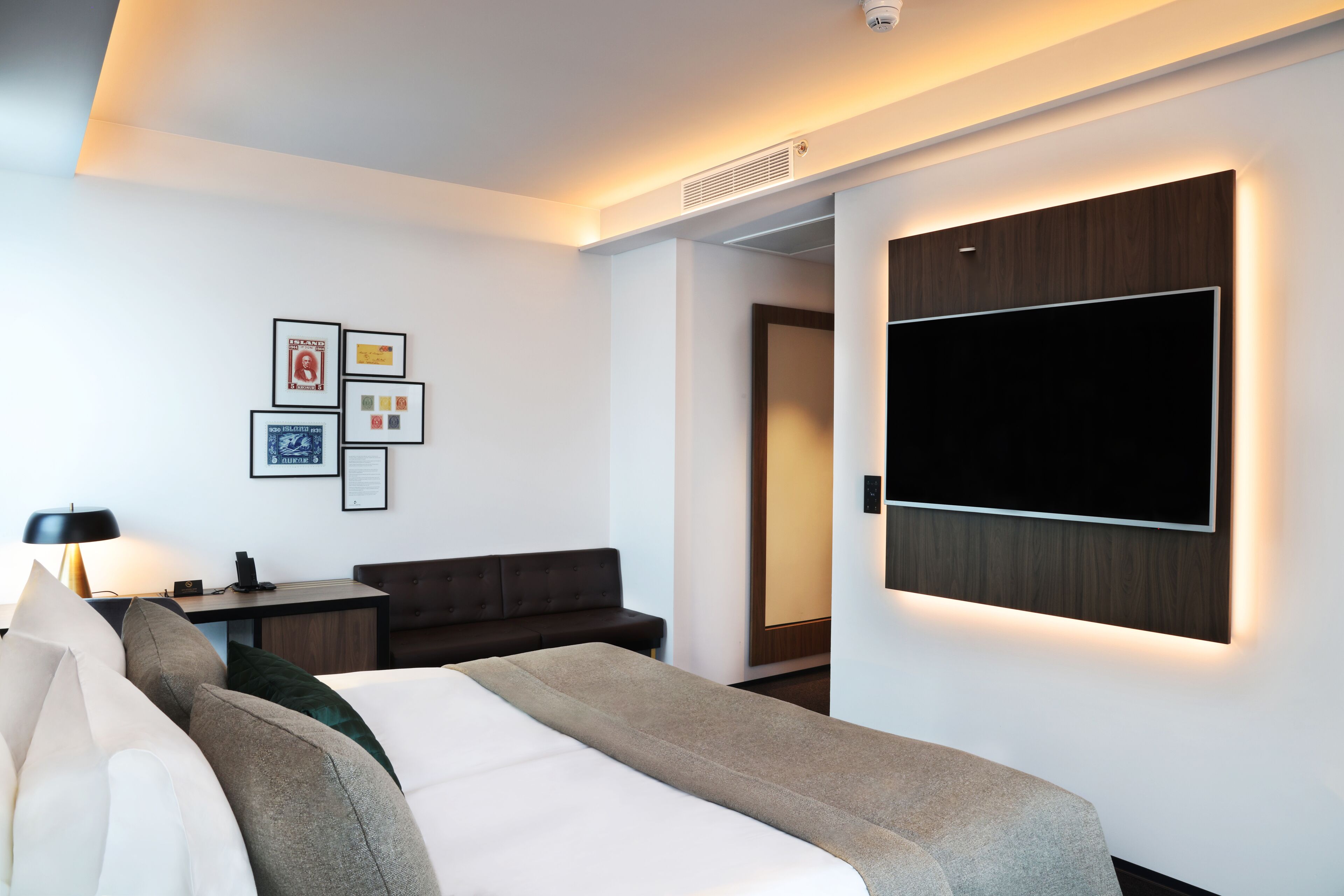 A modern hotel room with a bed, a desk, a sofa, and a TV mounted on the wall.