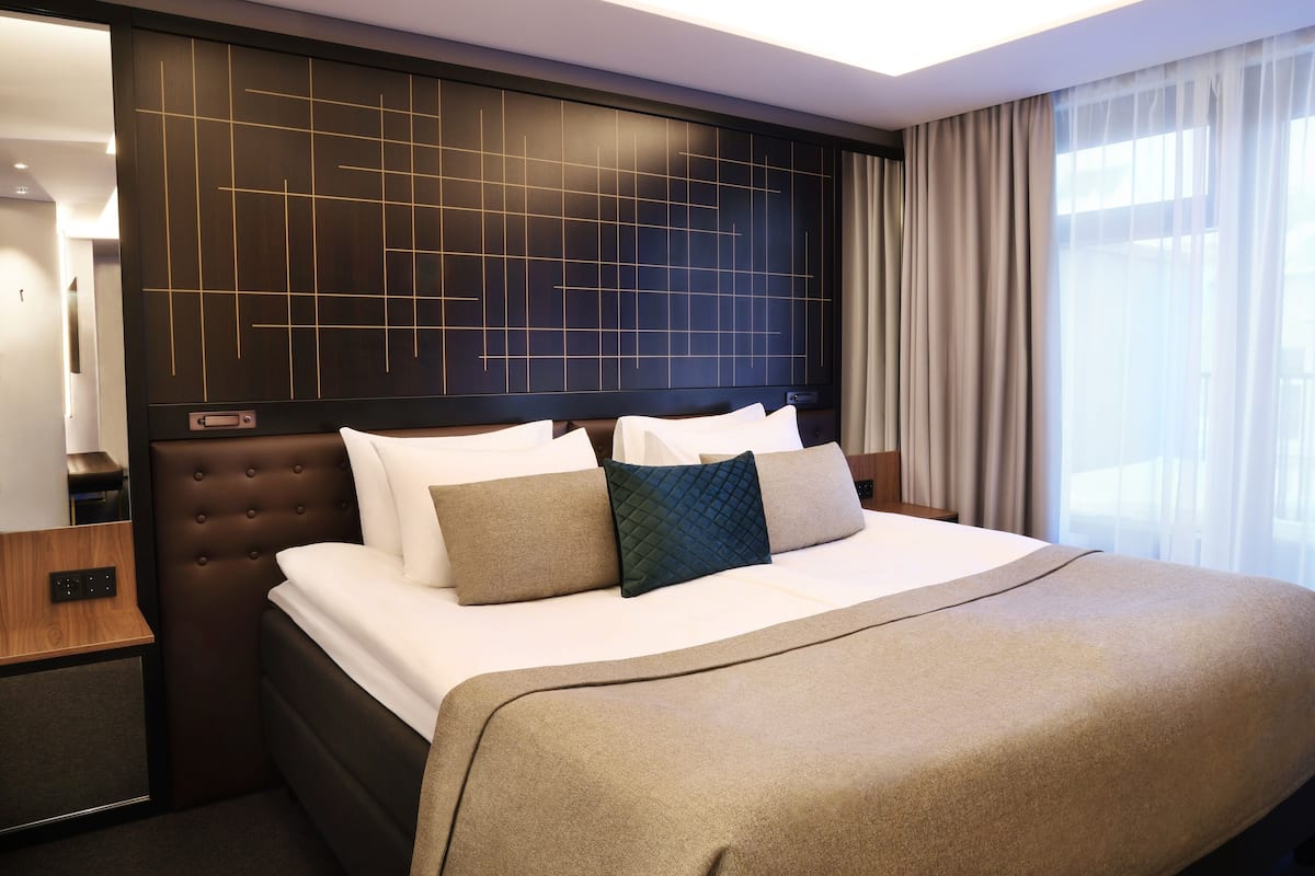 A hotel room with a large bed, a headboard with a geometric pattern, a bedside table, and a window with curtains.