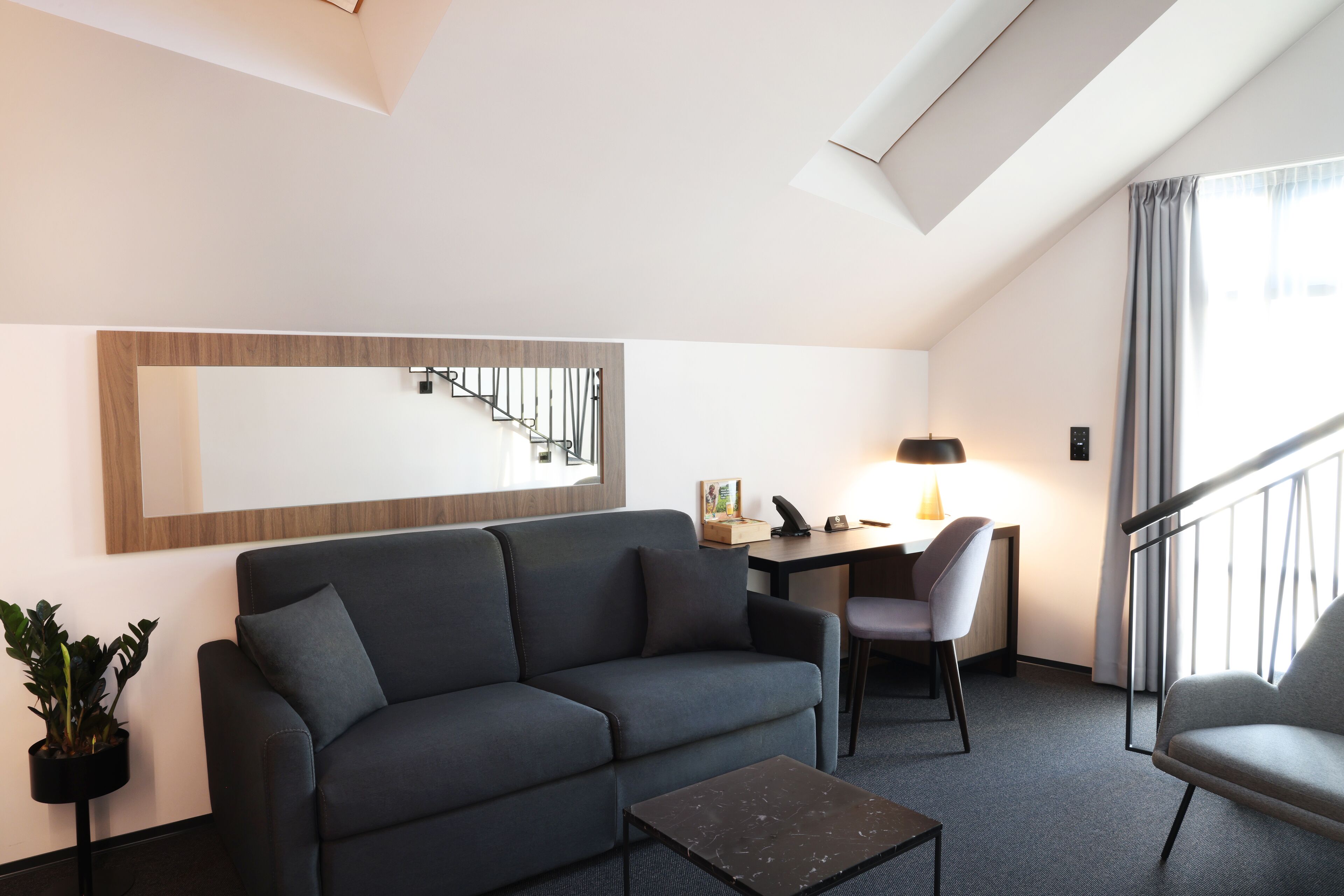 A modern hotel room with a grey sofa, a small coffee table, a desk with a chair, a potted plant, and a staircase.