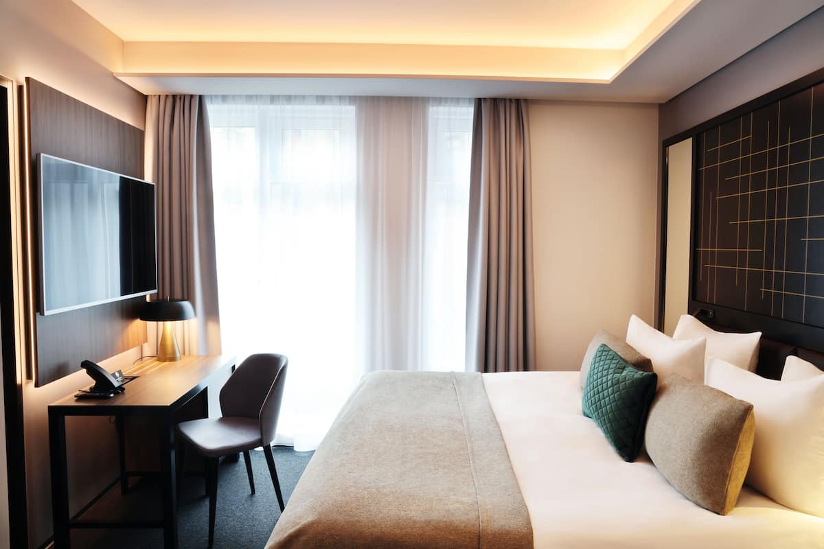 A hotel room with a bed, a desk, a chair, a TV, and a window with curtains.