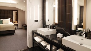 Una habitación de hotel moderna con una cama grande, dos lavamanos blancos, un tocador oscuro y un espejo amplio.