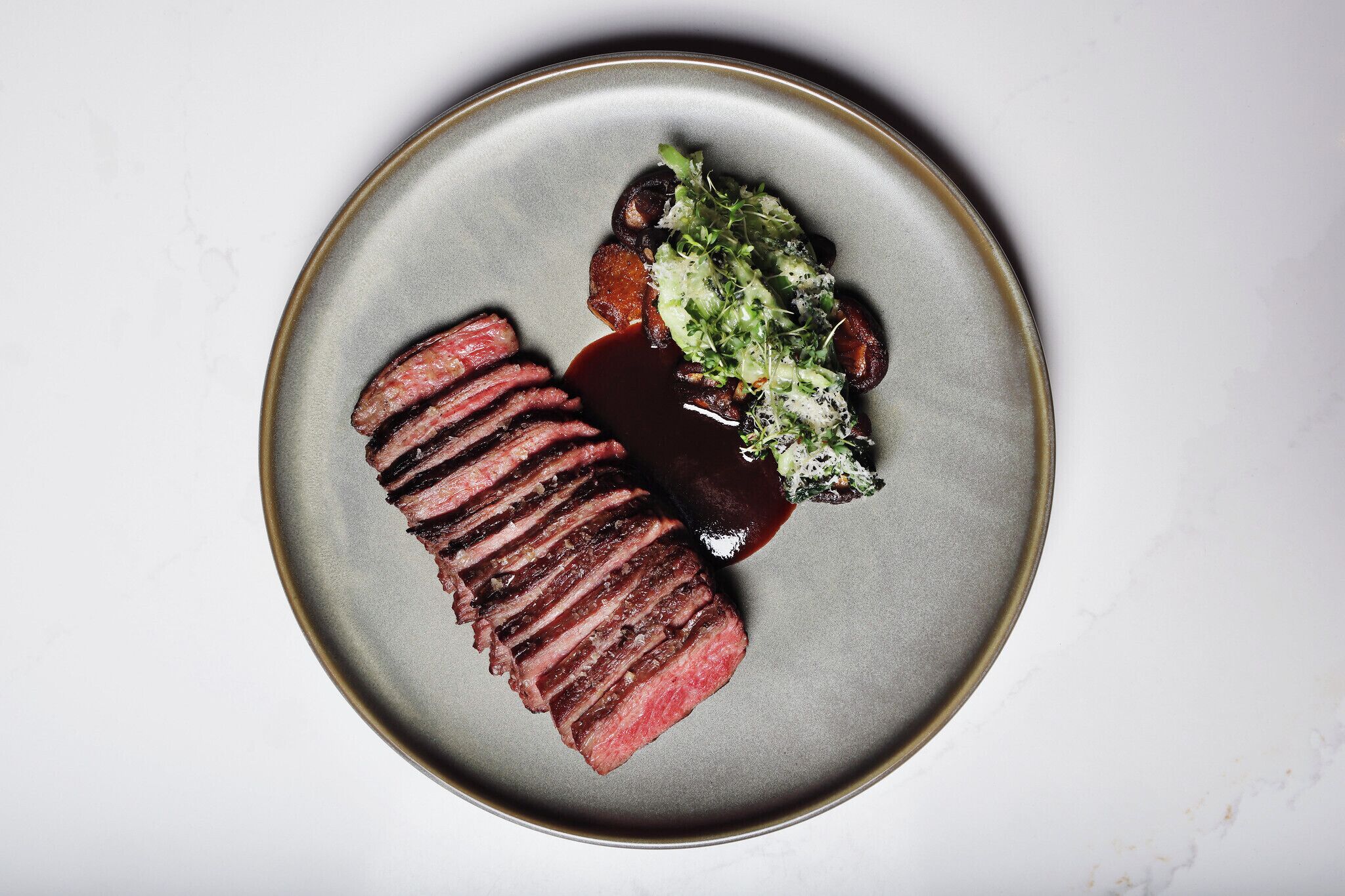 Un plato con bistec en rodajas, champiñones y una guarnición verde sobre un fondo de mármol blanco.
