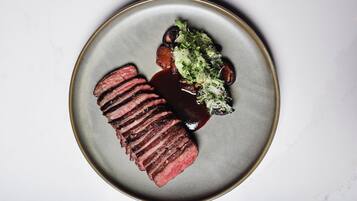 Un plato con bistec en rodajas, champiñones y una guarnición verde sobre un fondo de mármol blanco.
