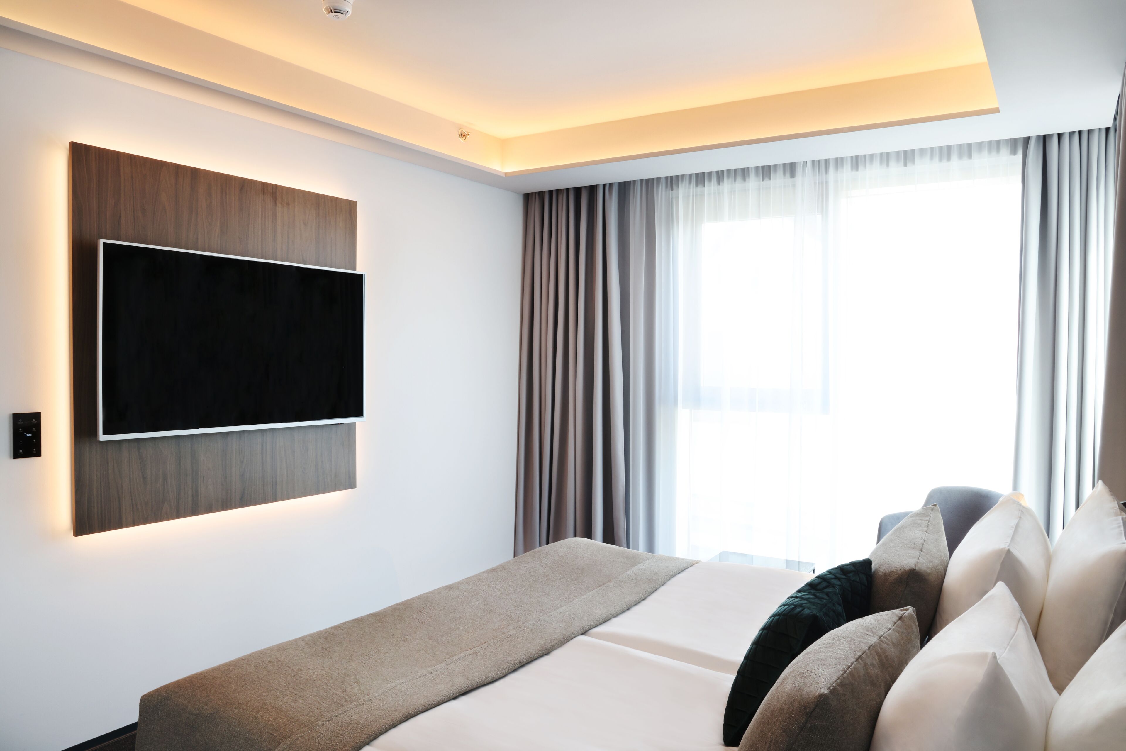 A modern hotel room with a large flat-screen TV mounted on the wall, a bed with a beige and white bedding, and a window with curtains.