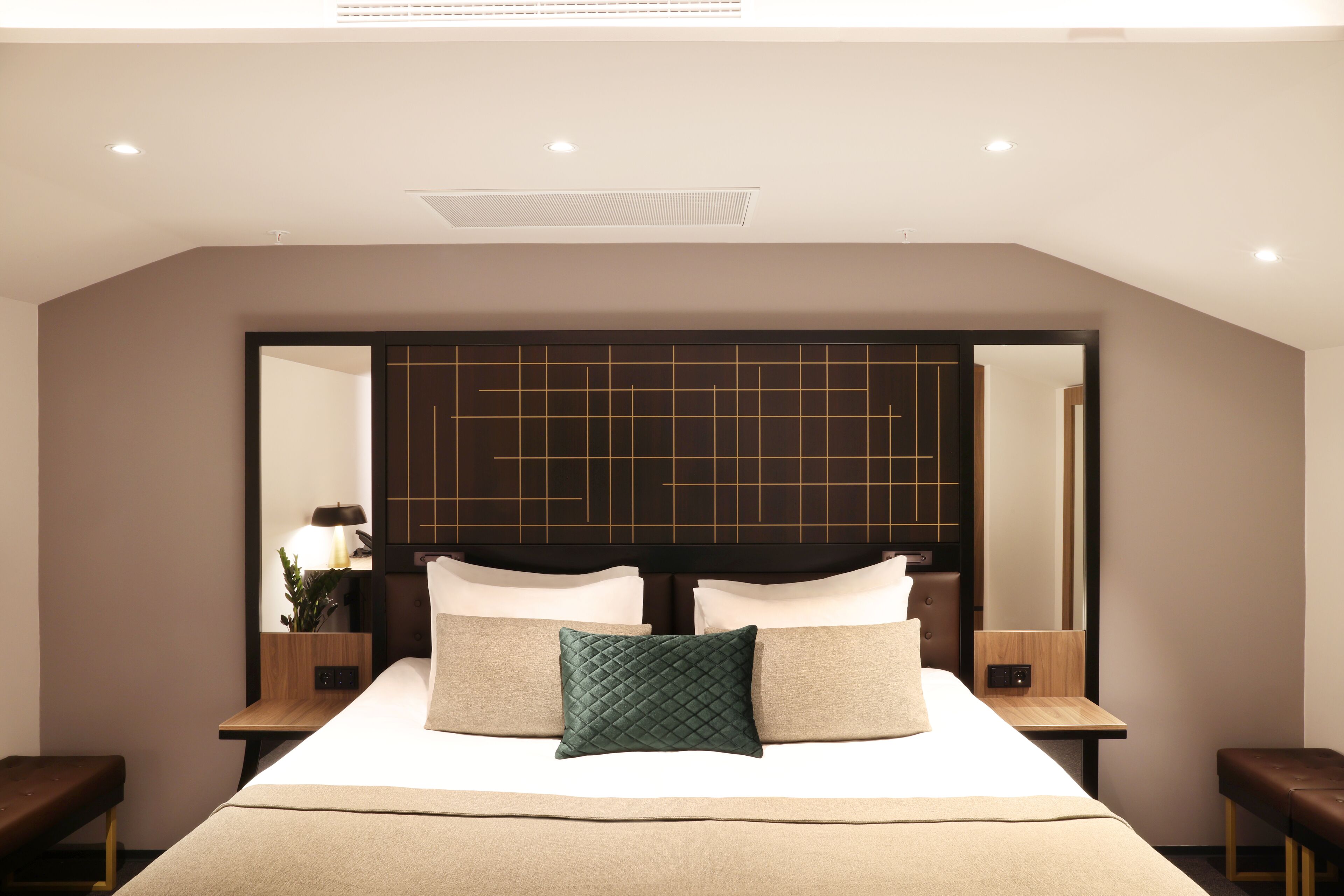 A modern hotel room with a large bed, a headboard with a grid pattern, two bedside tables with lamps, and a bench at the foot of the bed.