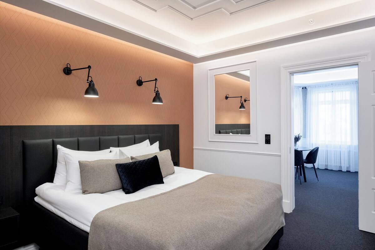 A modern hotel room with a large bed, a dark headboard, two wall-mounted reading lamps, a large mirror, and a view of the dining area through a door.