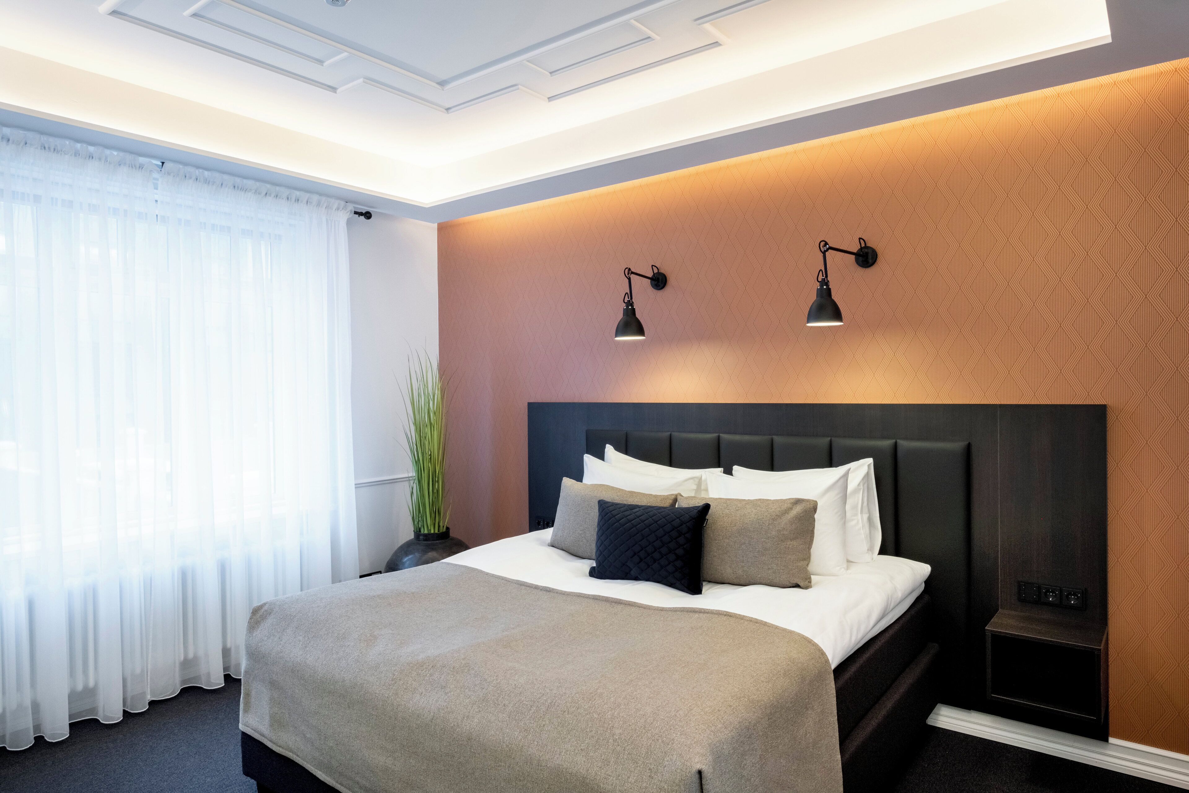 A hotel room with a large bed, a headboard, two wall-mounted lamps, and a window with sheer curtains.