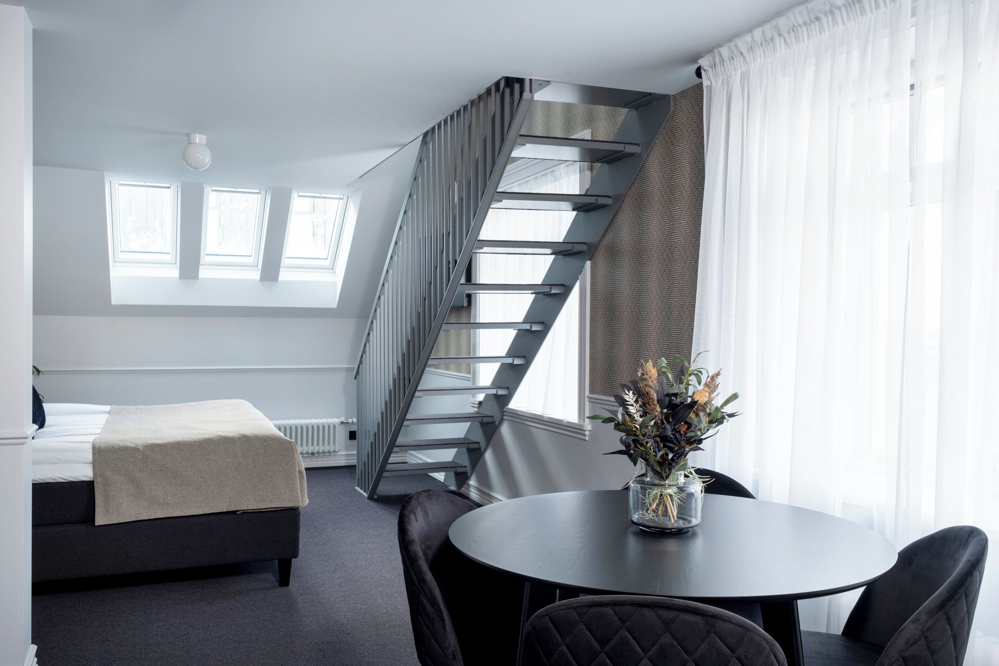 Une chambre moderne avec un lit, une table ronde entourée de chaises, un escalier et une fenêtre avec des rideaux.