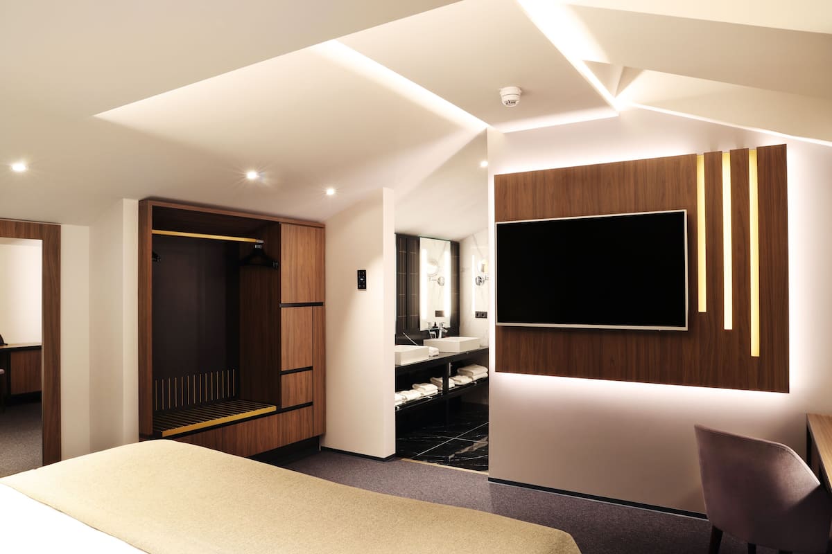 A modern hotel room with a large bed, a wardrobe, a vanity with a mirror, and a flat-screen TV mounted on the wall.