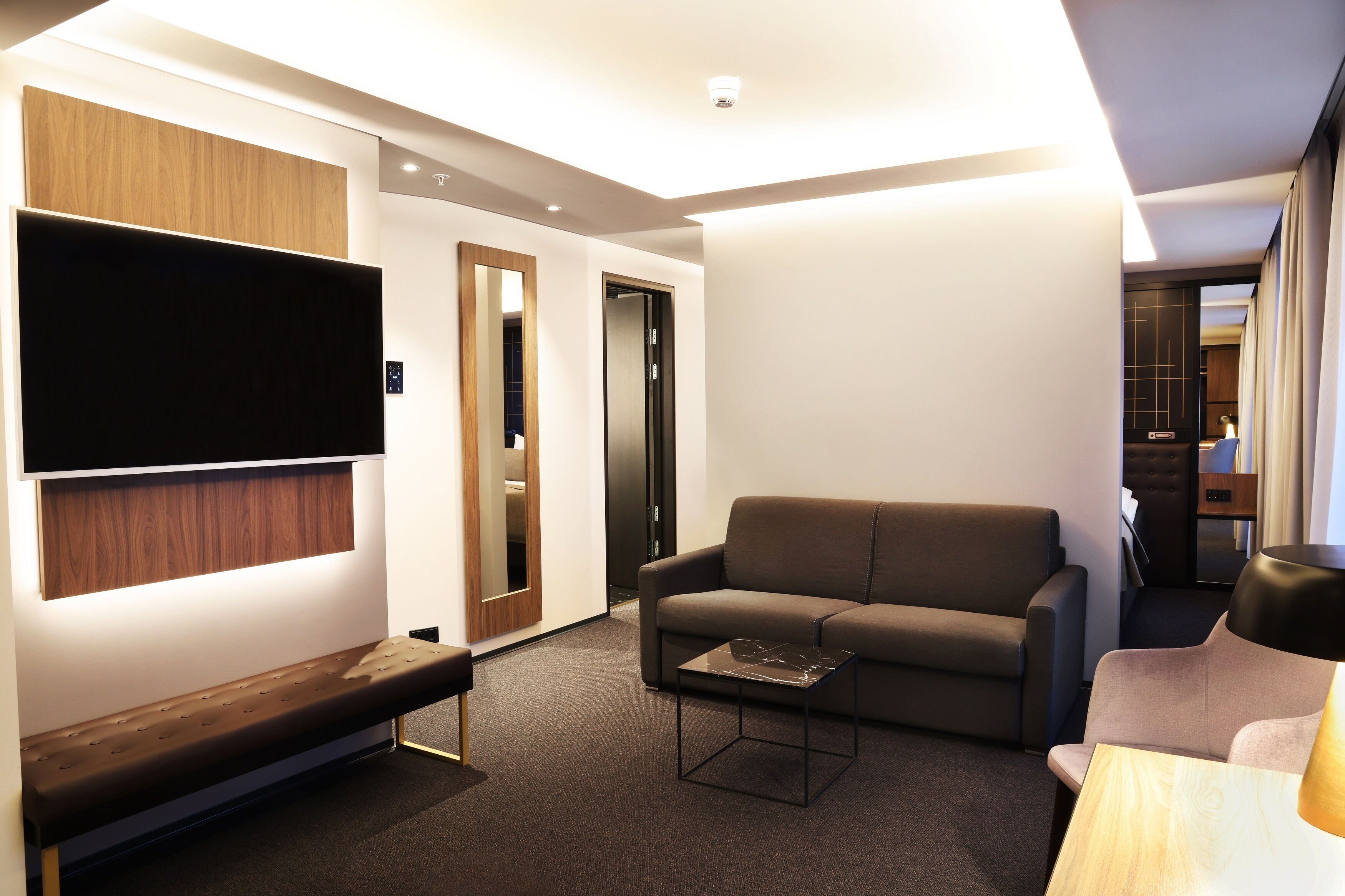 Uma sala de estar moderna com um sofá, um banquinho, uma mesa de centro, uma TV fixada na parede e uma porta que dá para outro cômodo.