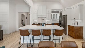 A modern kitchen with white cabinetry, a large island with bar stools, stainless steel appliances, and a wooden floor. - Gathering Place Luxury-Pool/Spa/OutdoorKitchen (Tulsa)