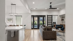 A modern kitchen with a central island, a dining area with a sofa and coffee table, and a view through a glass door to the outdoors. - Gathering Place Luxury-Pool/Spa/OutdoorKitchen (Tulsa)