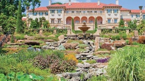 A large, multi-story building with a red-tiled roof, a central courtyard with a fountain, and a well-maintained garden with various plants and rocks. - Gathering Place Luxury-Pool/Spa/OutdoorKitchen (Tulsa)