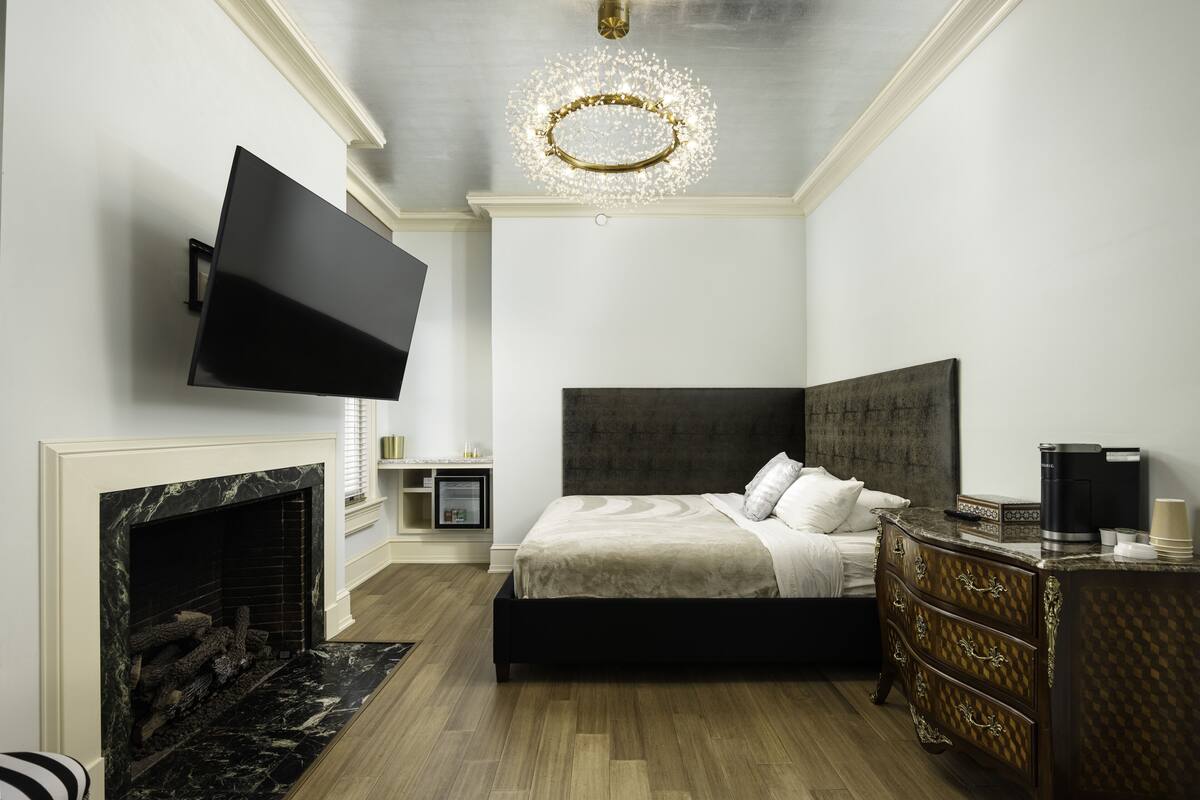 Un dormitorio moderno con un televisor de pantalla plana montado en la pared, una chimenea, una cama grande con cabecera oscura y un aparador de madera.