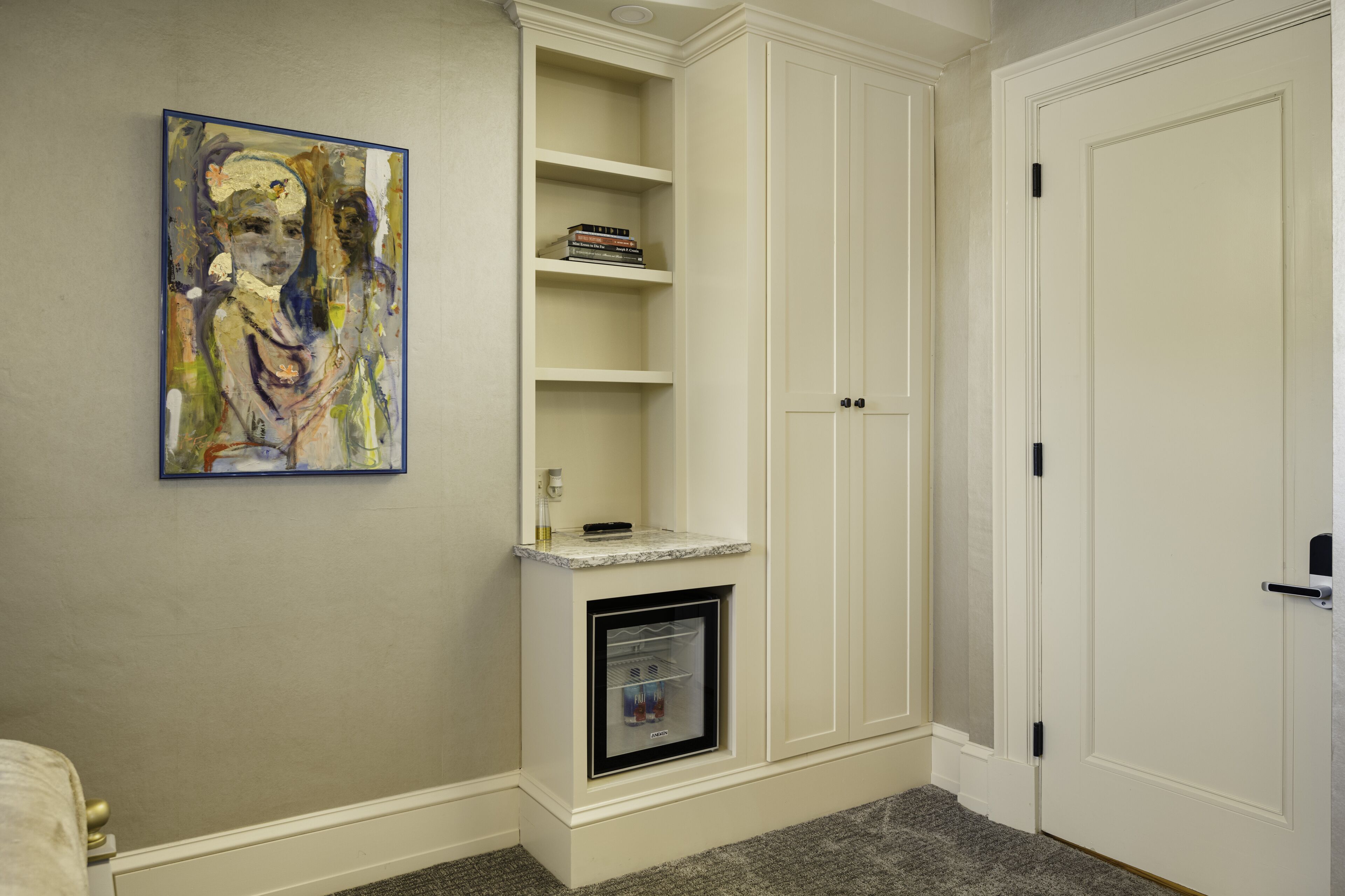 A corner room with a built-in mini-fridge, a shelf with books, and a painting on the wall.