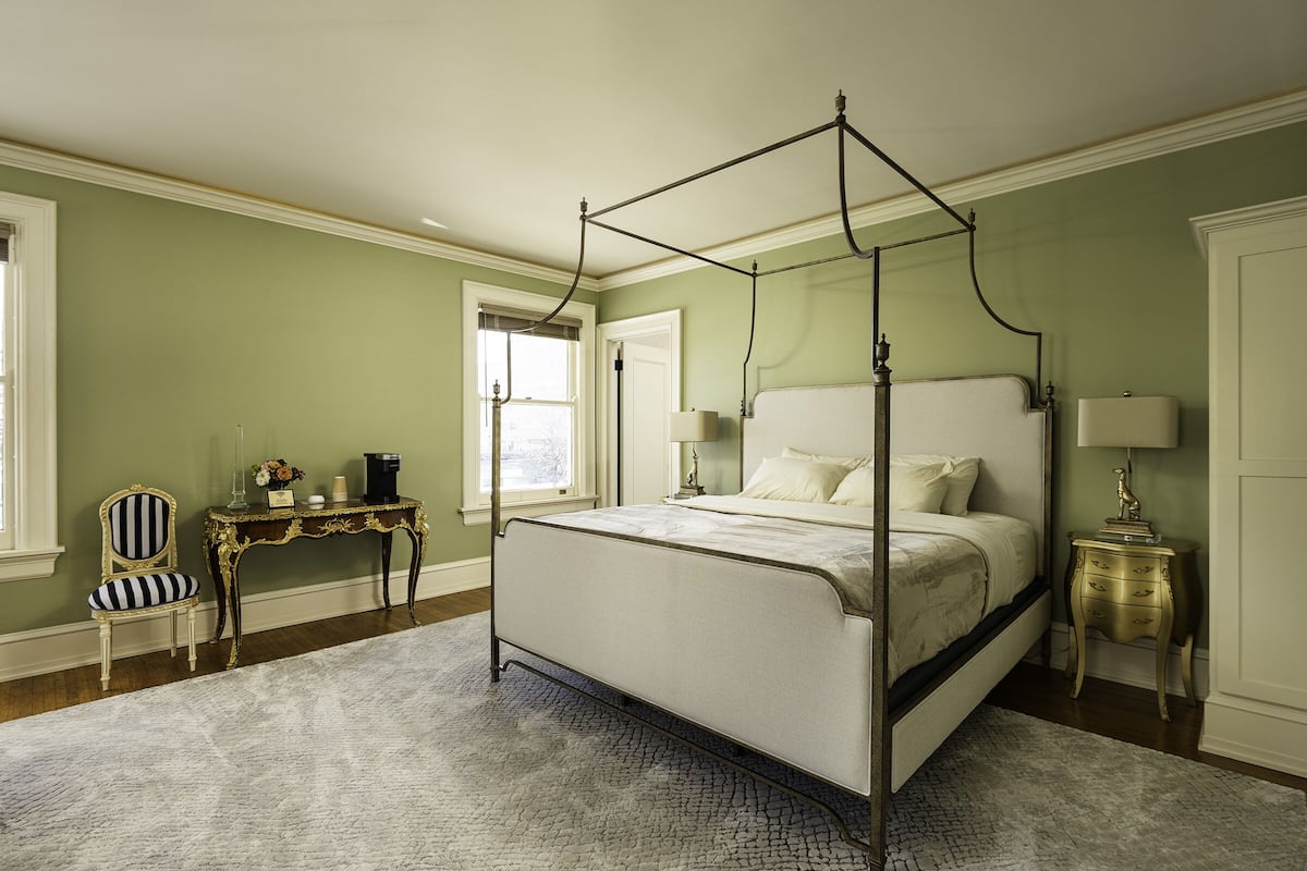 Una cama con dosel en habitación con paredes verdes, una silla blanca, una mesita y una mesita de noche.
