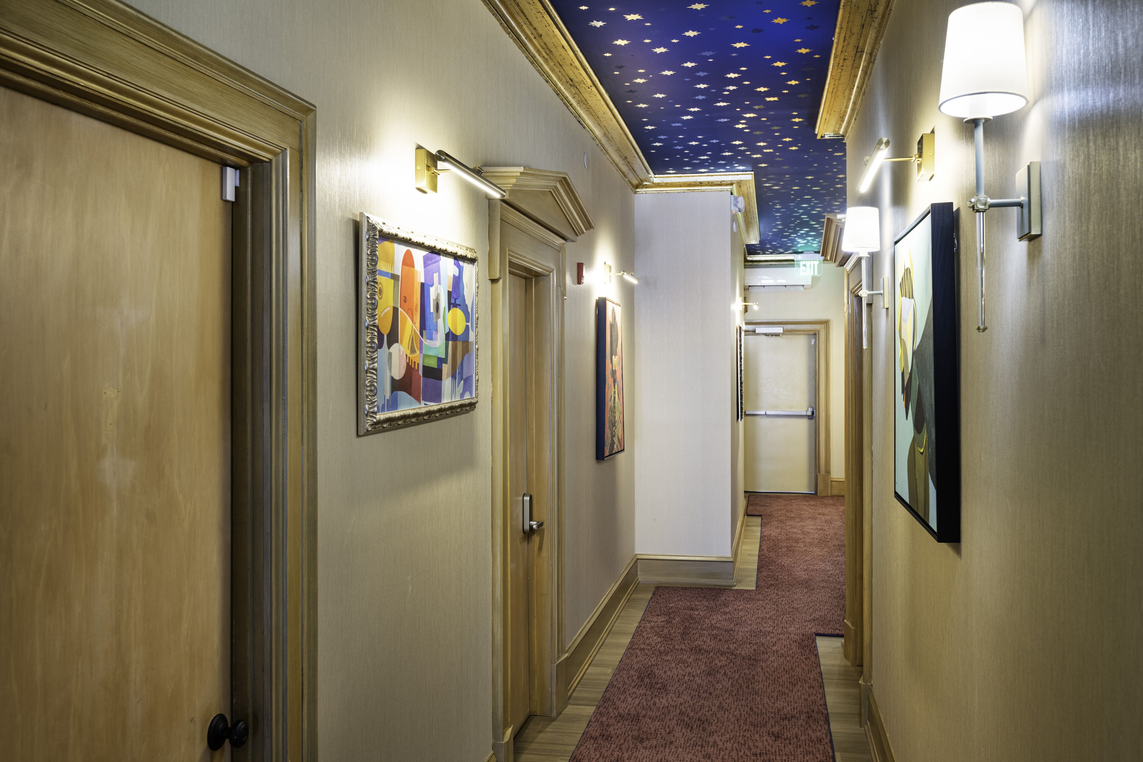 A hotel hallway with framed artwork on the walls, a carpeted floor, and a star-patterned ceiling.