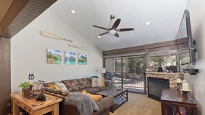 Un salon spacieux avec un grand canapé, une table basse en bois et une cheminée. On y trouve également un ventilateur de plafond, un téléviseur fixé au mur et une grande fenêtre offrant une vue sur l’extérieur.