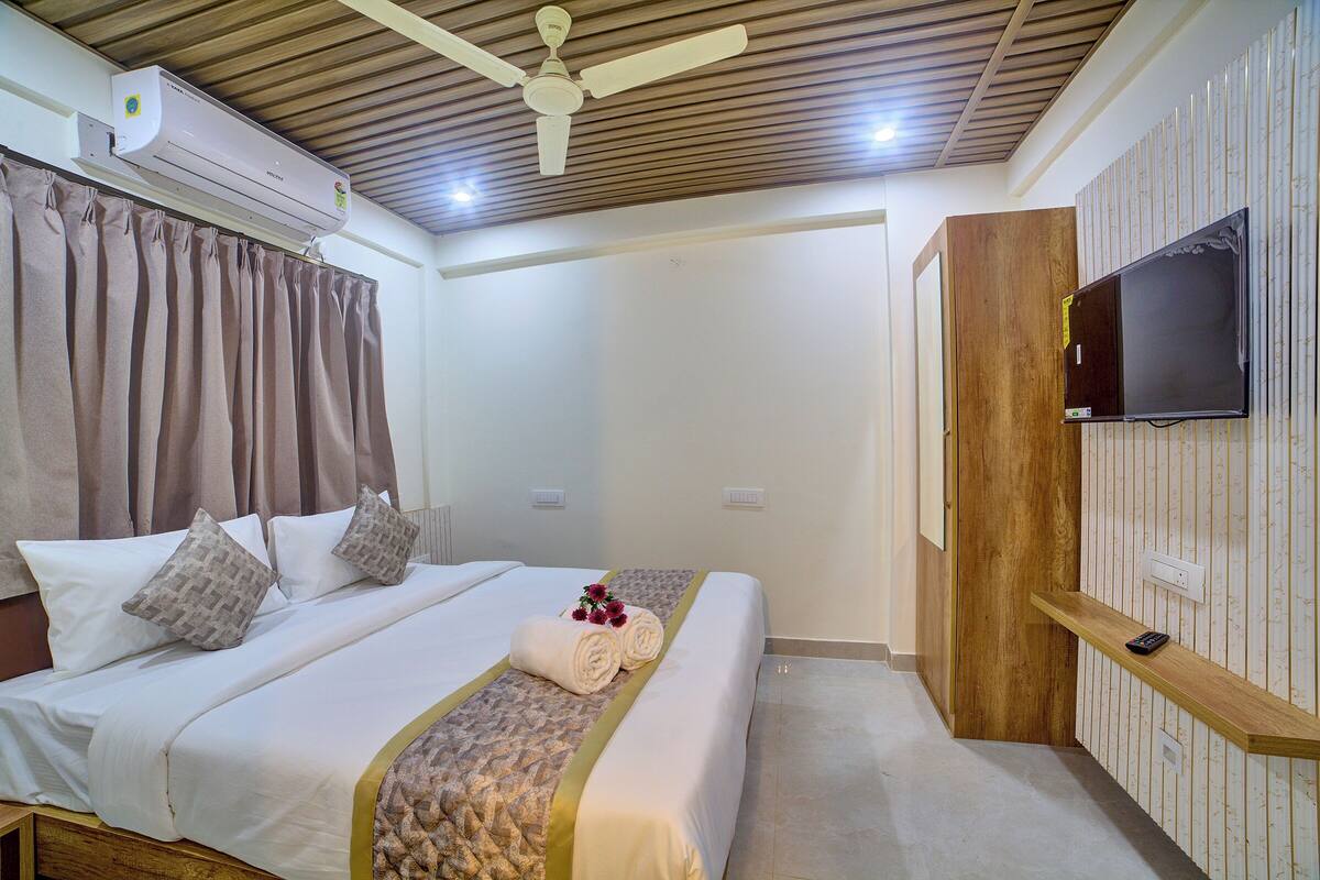 Une chambre d’hôtel avec un lit, des rideaux, un ventilateur de plafond, un climatiseur, une télévision et une étagère en bois.