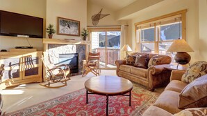 A cozy living room with a fireplace, comfortable seating, a flat-screen TV, and a large window with a mountain view. - Spacious 3-Bedroom Condo with Hot Tub, Surrounded by Beautiful Views - CS431 (Copper Mountain)