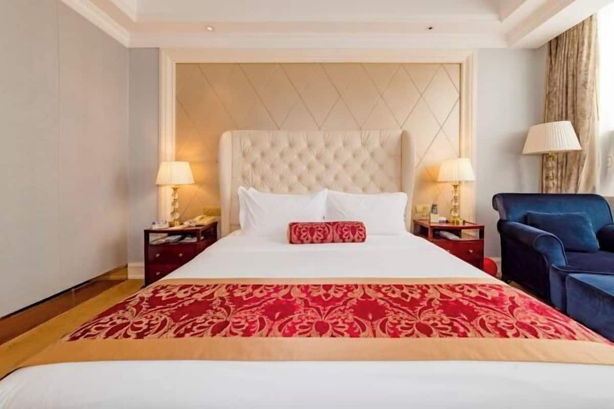 A hotel room with a large bed, two bedside tables with lamps, a blue armchair, and a patterned red and beige bedspread.