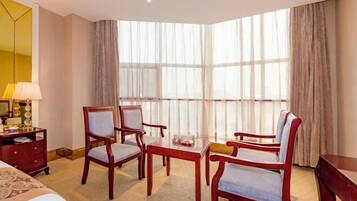 A hotel room with a bed, two armchairs, a small table, a lamp, a TV, and large windows with curtains.