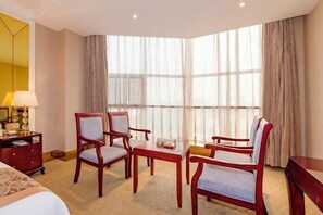 Une chambre d’hôtel avec un lit, deux fauteuils, une petite table, une lampe, un téléviseur et de grandes fenêtres avec des rideaux.