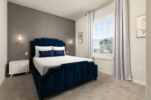 Une chambre à coucher avec un lit recouvert d’un tissu bleu foncé, du linge de lit blanc, une table de chevet avec des tiroirs, un luminaire fixé au mur et une fenêtre avec des rideaux blancs.
