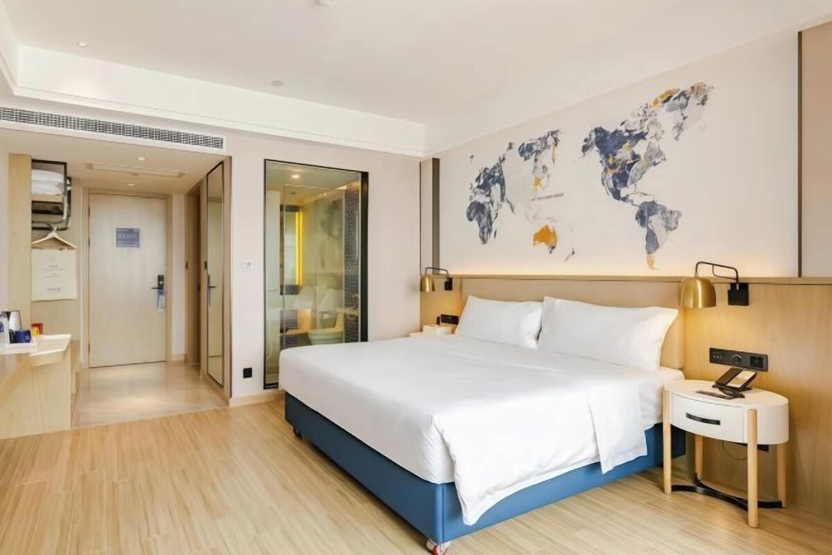 Une chambre d’hôtel avec un grand lit, une table de chevet avec une lampe, une carte du monde accrochée au mur et une porte donnant sur une salle de bain.