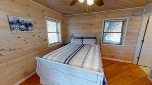 Ein Schlafzimmer mit Holzwänden, einem Bett, zwei Fenstern und einem Deckenventilator.