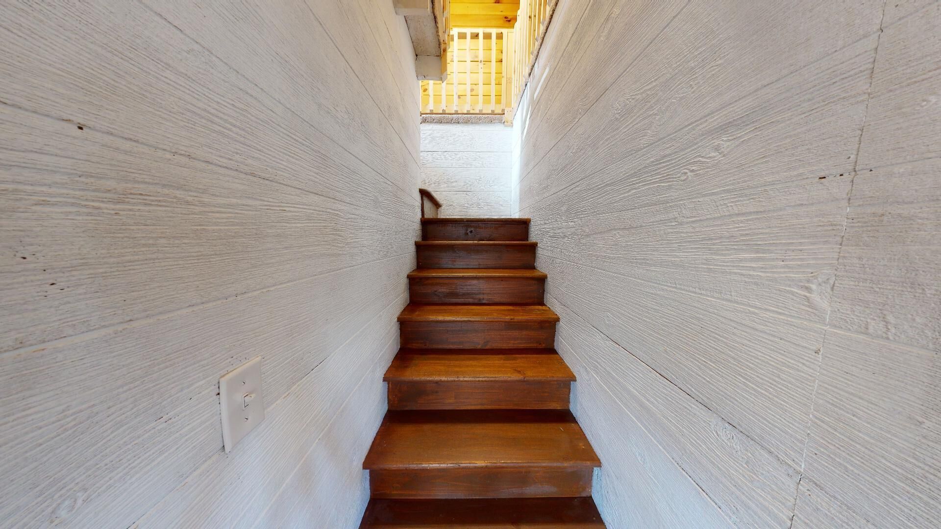 Un escalier en bois, avec des murs de couleur claire de part et d’autre, mène à une pièce lumineuse située à l’étage supérieur.