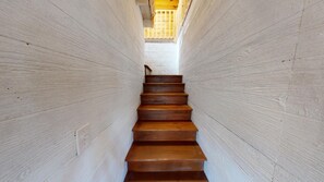 A wooden staircase with a light-colored wall on either side, leading to a bright room at the top.