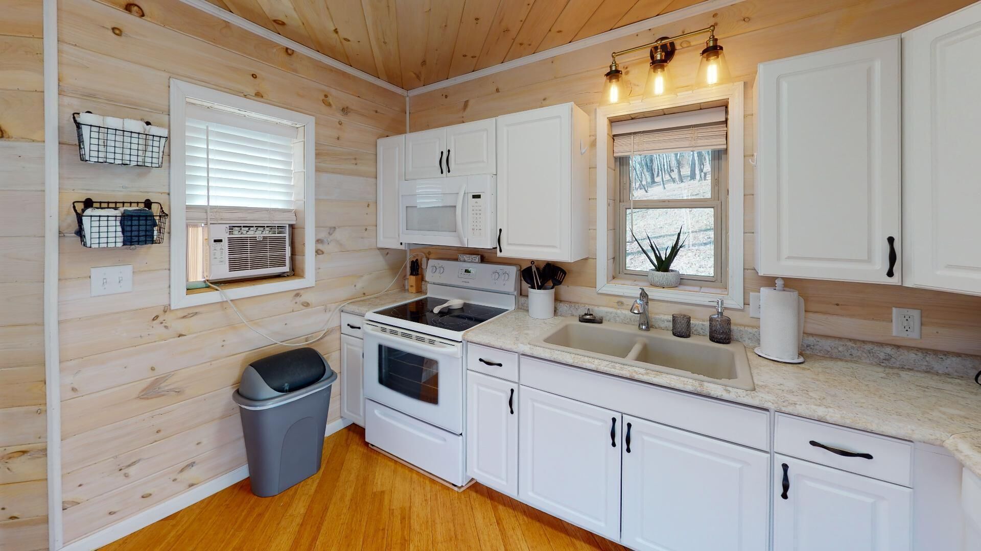 A compact kitchen with white cabinets, a microwave, stove, and a sink. There is a window with blinds and a small trash bin.