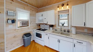 A compact kitchen with white cabinets, a microwave, stove, and a sink. There is a window with blinds and a small trash bin. - Luxury Cottage in Nelsonville near Hocking Hills (Nelsonville)