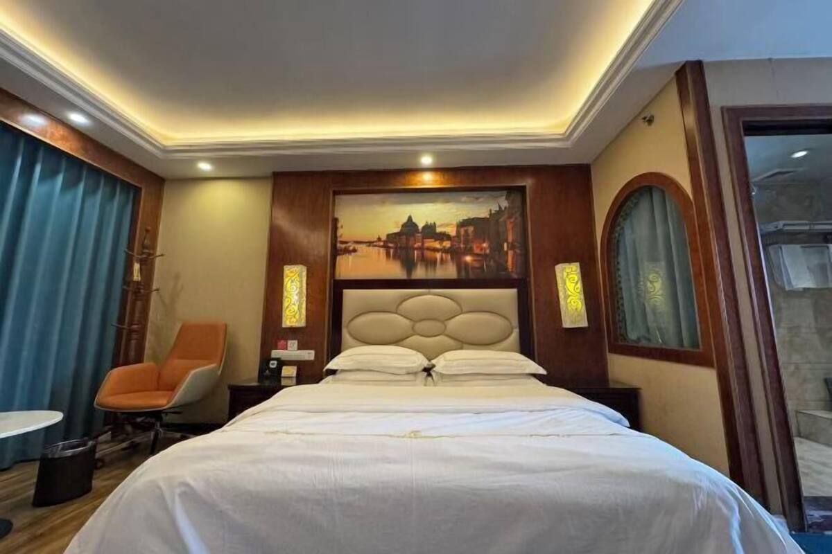 A hotel room with a large bed, a chair, a painting, and a view of a cityscape.